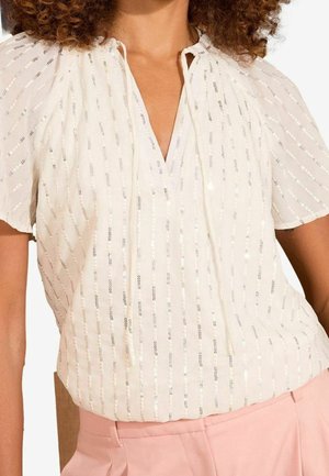 Kvinde iført hvid bluse med korte ærmer og lodrette sølvglitrende pailletter, kombineret med højtaljede lyserøde bukser, vist fra hals til hofter.