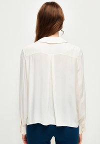 Blusa de crema claro con detalle de pliegue en la parte posterior, con cuello y puños abotonados. Tejido suave con un ajuste relajado.