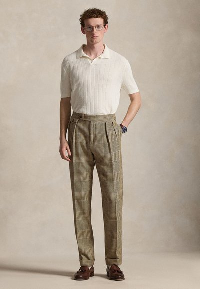 Άνδρας με σγουρά μαλλιά και γυαλιά φορώντας πλεκτό πόλο σε εκρού απόχρωση, καρό παντελόνι ψηλόμεσο, καφέ μοκασίνια και ρολόι χειρός, όρθιος μπροστά σε μπεζ φόντο.