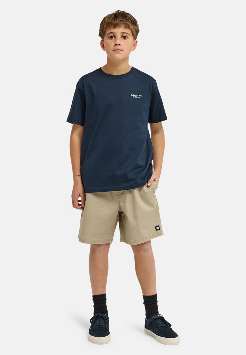 Garçon debout, mains dans les poches, portant un t-shirt bleu marine, un short beige, des chaussettes noires et des baskets bleu marine sur fond blanc.