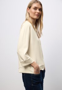 Top de color crema, de manga larga, con cuello con cremallera y corte holgado. Presenta una tela suave y lisa, con puños acanalados, combinado con jeans oscuros.