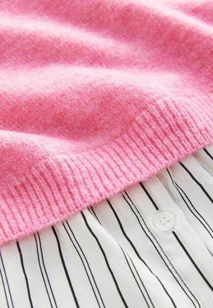 Růžový pletený svetr s žebrovaným lemem položený na bílé košili s knoflíky, která má černé vertikální pruhy. Měkká textura a vrstvený design.