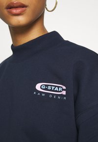 Marinblå sweatshirt med ribbad krage och en logotyp på framsidan i rosa och blått, tillverkad av mjukt material, med en avslappnad passform.