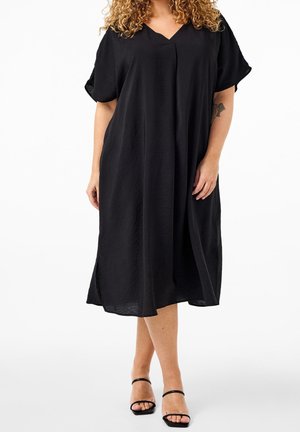 Kvinde med krøllet hår iført en løs sort midi-kjole med korte ærmer og sorte åbne sandaler mod en hvid baggrund.