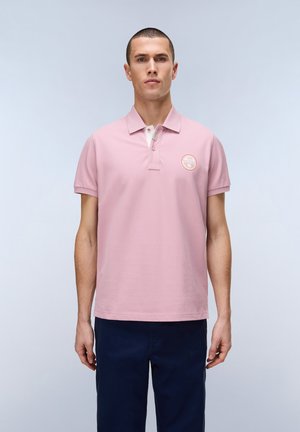 Mann mit kurzen Haaren, der ein hellrosa Poloshirt mit Logo auf der Brust und dunkelblaue Hose trägt, steht vor einem schlichten hellen Hintergrund.