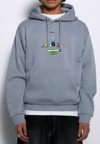 Jasnoszara bluza z kapturem z miękkiego materiału, z kieszenią z przodu. Grafika w pikselach przedstawia postać i różne ikony, podpisane "LOKALNI BOHATEROWIE GRAJĄ".