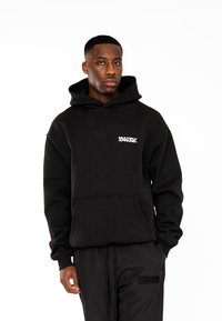 Sudadera negra con bolsillo frontal, que presenta un logotipo blanco "MULTIPLY APPAREL". Está fabricada con una tela suave, con un corte relajado y capucha ajustable.