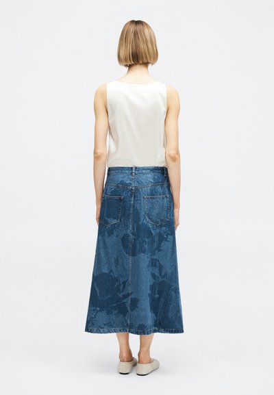 Γυναίκα που φαίνεται από πίσω, φοράει λευκό αμάνικο μπλουζάκι, μπλε τζιν φούστα μέχρι τους αστραγάλους με πίσω τσέπες και ανοιχτόχρωμα επίπεδα παπούτσια.