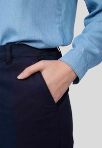 Chemise en denim bleu avec un col, associée à un pantalon bleu marine foncé ; la main repose sur la poche du pantalon, montrant un design simple et fonctionnel.