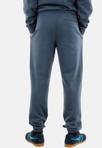 Blaue Jogginghosen mit elastischen Bündchen, Seitentaschen und einer Gesäßtasche. Weicher Stoff mit glatter Textur. Kombiniert mit dunklen Schuhen mit blauen Akzenten.