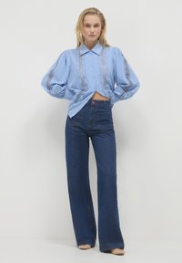 Blouse bleu clair avec des manches bouffantes et des détails en dentelle élaborés, associée à un jean flare taille haute bleu foncé et des chaussures nudes.