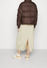 Braune Pufferjacke, beiger Rock mit Schlitz und cremefarbene Sneaker. Zu den Merkmalen gehören elastische Bündchen und ein verstellbarer Rockzug.