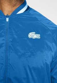 Blå bomberjacka med en slät, glänsande textur, med en vit och blå randig krage, hel dragkedja och en broderad logotyp.