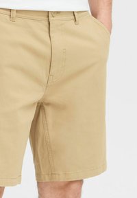 Shorts en coton beige avec un devant plat, des poches latérales et une fermeture à bouton. Le tissu semble lisse avec des détails de coutures visibles.