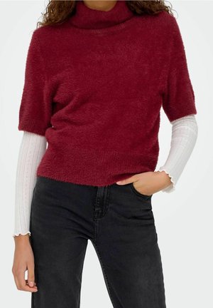 Mujer con un suéter rojo y peludo de manga corta sobre una camiseta blanca de manga larga combinado con vaqueros negros de cintura alta, mano en el bolsillo.