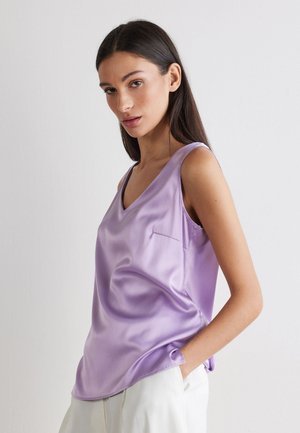 Femme portant un haut en satin lavande sans manches avec col en V et un pantalon blanc, posant avec une main dans la poche devant un fond uni.