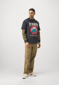 Marine blå grafisk t-skjorte med teksten "SUMMER LEAGUE" og Nike-logo, lagd over en stripete langermet skjorte; beige bukser og hvite sneakers.
