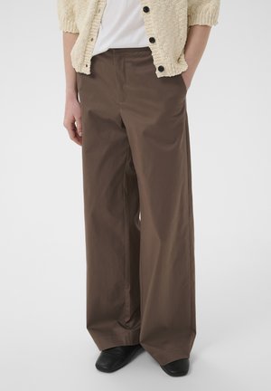 Personne portant un pantalon large marron ample, des chaussures noires, une chemise blanche et un cardigan en tricot beige boutonné, les mains dans les poches.