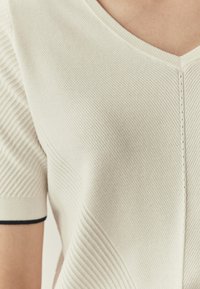 Haut en tricot couleur crème avec un col en V, une texture côtelée, des manches courtes avec un accent noir à l'ourlet et des détails de coutures subtils.