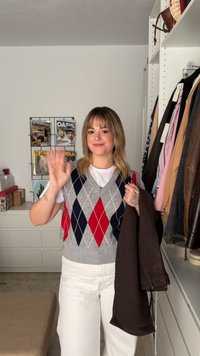 Šedý argyle sveter s červenými a námorníckymi diamantmi, biele tričko a hnedá bunda. Svetlé široké nohavice. Uložené pozadie s visiaciami odevmi.