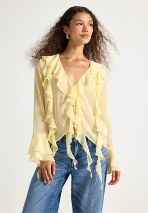 Helle gelbe, transparente Bluse mit Rüschendetails entlang des Ausschnitts und der Ärmel. Mit einer Schleife vorne, kombiniert mit lockeren Jeans.