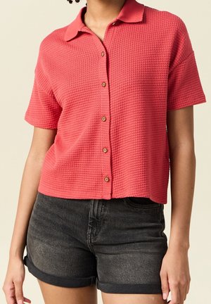 Vrouw draagt een gestructureerd koraalkleurig kort mouwen overhemd met knopen en zwarte denim shorts met opgerolde zoom, staand tegen een effen achtergrond.