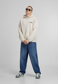 Krémová mikina s logem "PEOUS", klokaní kapsa, oversized střih; kombinována s volnými modrými džíny a sneakers s zelenými detaily.