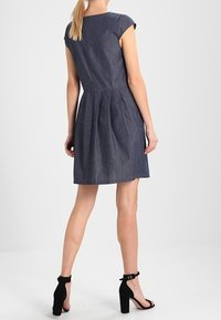 Denimkleid mit kurzen Ärmeln, gefaltetem Rücken und taillierter Taille. Dunkelblaue Farbe. Getragen mit schwarzen High Heels.