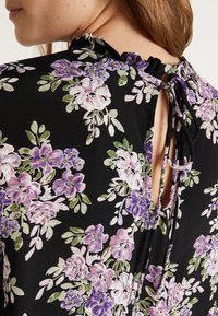 Sort blomsterbluse med lilla og grønne mønstre, der har en flæset krave og en snøre lukning bagpå, som viser en åben rygdetalje.