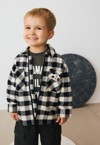 Veste en flanelle à carreaux noir et blanc avec un visage de panda sur la poche, portée sur une chemise grise, associée à un pantalon cargo noir.