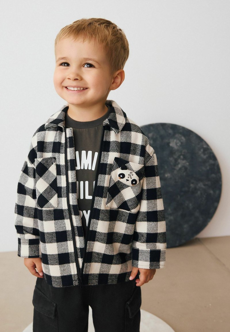Veste en flanelle à carreaux noir et blanc avec un visage de panda sur la poche, portée sur une chemise grise, associée à un pantalon cargo noir.