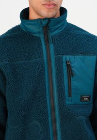 Uomo che indossa una giacca di pile blu teal con colletto rialzato, zip frontale completa e tasca sul petto con zip e etichetta del marchio.