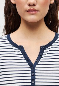 Ριγέ πουκάμισο henley με ύφασμα από βαμβάκι σε ναυτικό μπλε και λευκό, λαιμόκοψη σε σχήμα V και τέσσερα ναυτικά μπλε κουμπιά κατά μήκος της πατιλέτας.