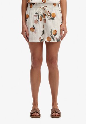 Femme portant un short blanc avec motif de fruits oranges, taille élastique avec cordon, et sandales plates marron, debout sur fond blanc.