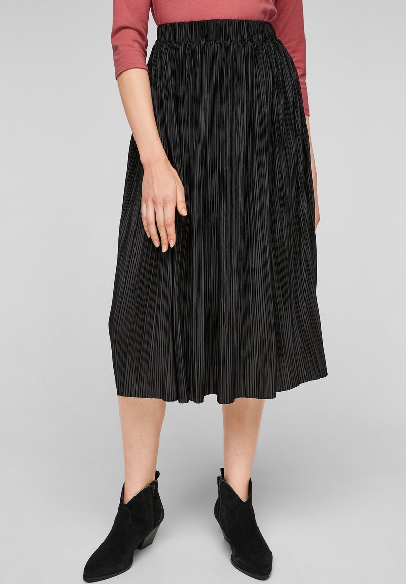Femme portant une jupe midi plissée noire, des bottines noires et un haut à manches longues rose poudré, debout sur un fond gris clair.