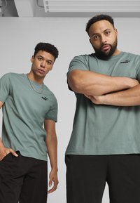 Dva muži mají na sobě volné světle zelené bavlněné trička s černým logem Puma na hrudi. Jsou stylizováni s černými kalhotami.