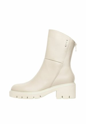 Stivale in pelle color panna fino al polpaccio con suola chunky, tacco a blocco, cerniera laterale e piccolo logo metallico vicino al bordo superiore.