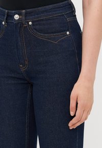 Close-up of dark blue denim jeans with visible stitching and silver rivets, worn by a person with a relaxed hand at their side.