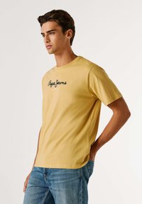 Camiseta de algodón amarilla con escote redondo, mangas cortas y un logotipo negro que dice "Pepe Jeans London" en el frente. Presenta un corte relajado.