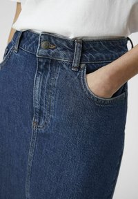 Denimskjørt i mørk blått, med høy midje, frontlomme, og kontrasterende sømmer. Metal knapp i midjen for lukking.