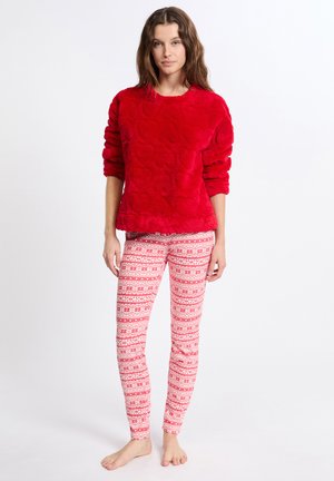 Rode pluizige trui met een getextureerd hartpatroon, gecombineerd met strakke witte leggings met rode en roze geometrische ontwerpen, op blote voeten.
