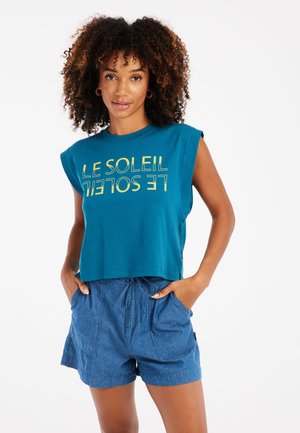 Teal kort mouwloos topje met gedurfde gele letters, gecombineerd met blauwe shorts met trekkoord, wat een casual en comfortabele stijl benadrukt.