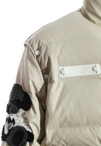 Chaqueta acolchada en beige con un acabado suave y brillante. Presenta botones a presión y un parche texturizado en negro y gris en la manga izquierda.