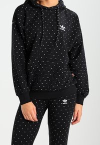 Person i svart Adidas-hoodie och leggings med små vita triangelmönster och Adidas-loggor på bröstet och låret.