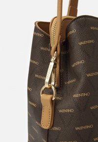 Acercamiento de un bolso marr�ón de Valentino con cuero texturizado, cremallera dorada y cierre de oro grabado con el logo, adjunto a una correa de cuero marrón.