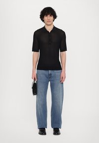 Fekete bordázott rövid ujjú pólóing gombos kivágással, laza világoskék farmernadrággal és fekete cipővel párosítva, valamint egy kis táskával a kezében.