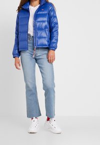 Modrá péřová bunda s lesklým povrchem, na zip, elastickými manžetami a logem na levé části hrudi. Kombinováno se světle modr ými džíny a bílými teniskami.