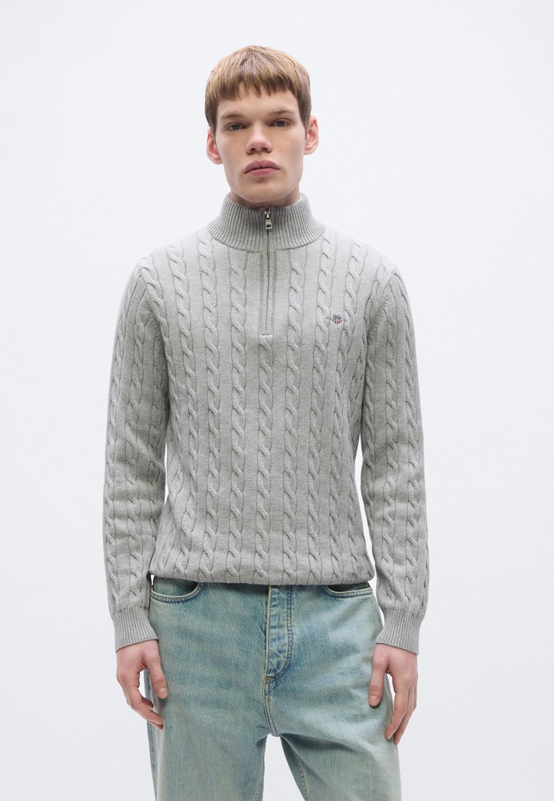 Γκρι πουλόβερ με πλέξη καλσόν, ψηλό γιακά και φερμουάρ μέχρι το στήθος. Διαθέτει ριμπ μανσέτες και τελείωμα· μικρό λογότυπο στο αριστερό στήθος. Φοριέται με ανοιχτόχρωμο τζιν.