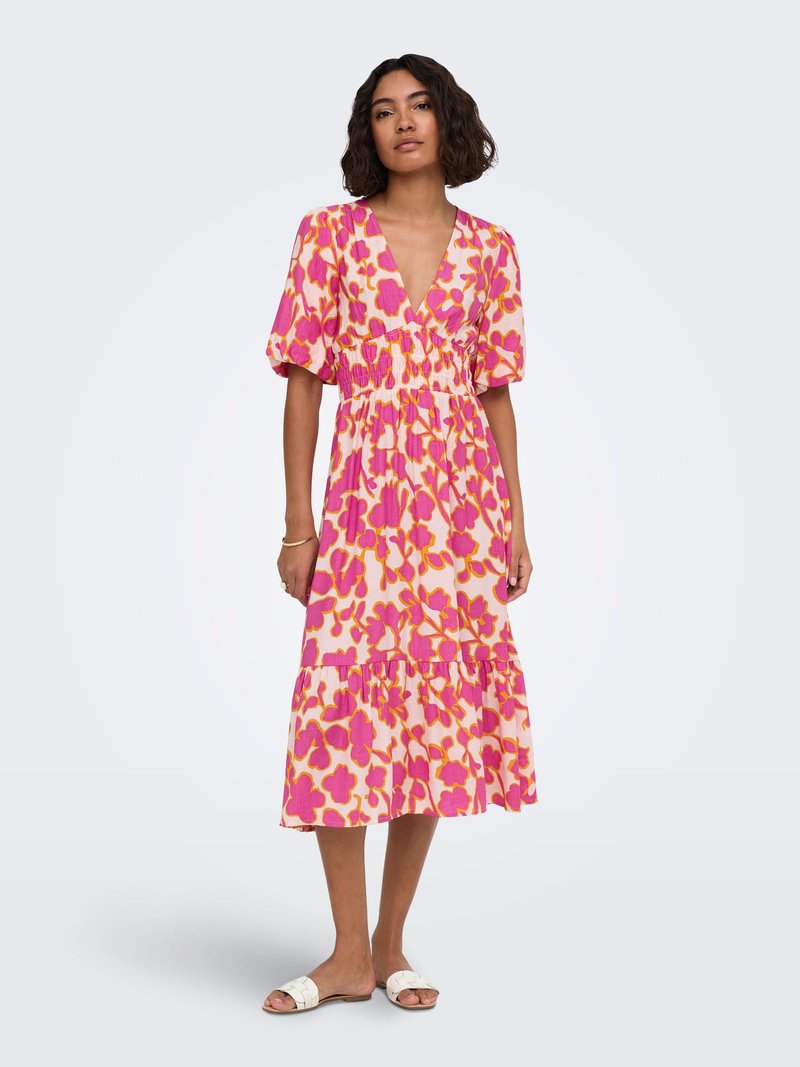 Femme portant une robe midi rose avec des motifs floraux orange et violet, manches bouffantes, col en V et sandales blanches à enfiler.