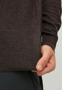 Pull marron foncé en tissu doux, avec poignets et ourlet côtelés, et une petite étiquette de marque sur le côté. Design texturé et décontracté.